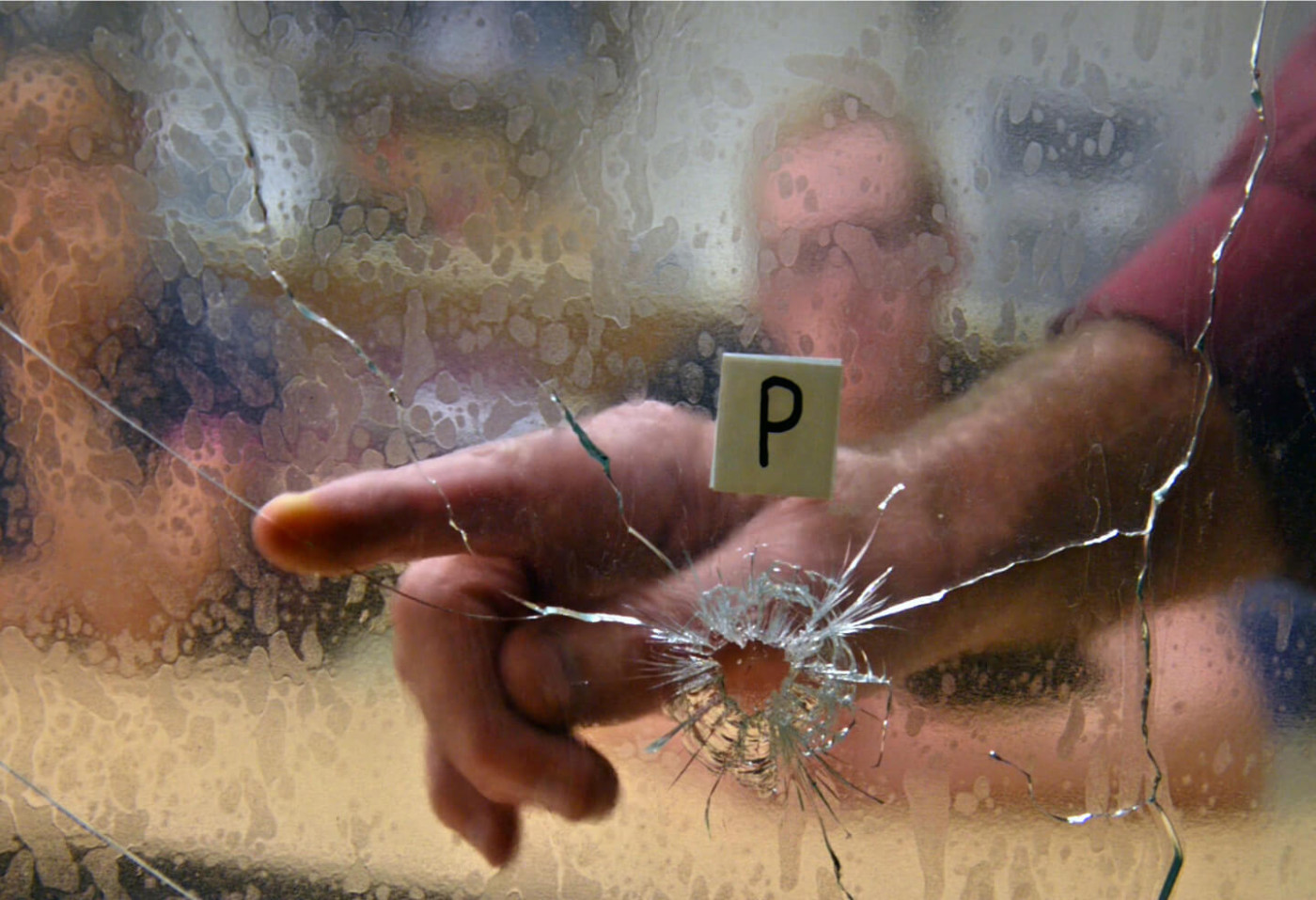 a forensics scientist is examining a bullet hole in glass as evidence whiile two people are observing from behind a table.