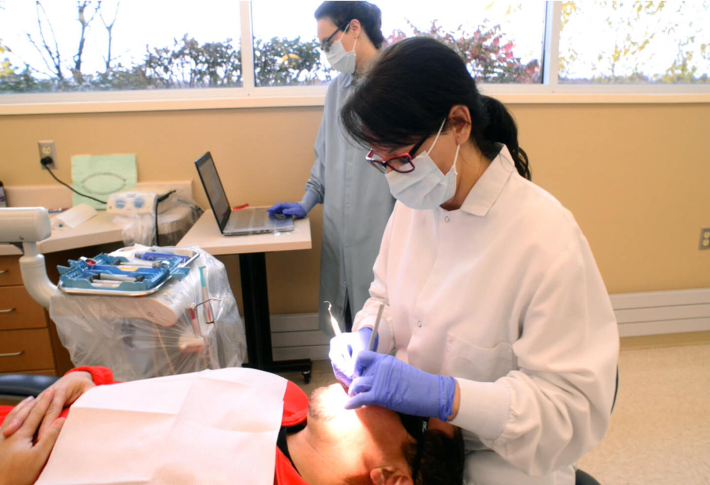 A Dental Therapist is working on the teeth of a patient, while another dental staff person is looking at records on a laptop. 