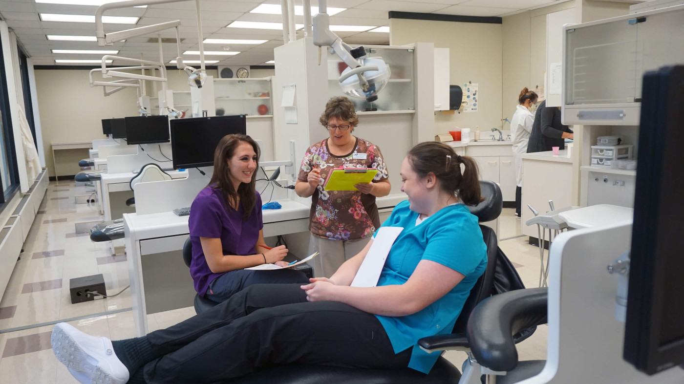 A Dental Assistant in-training is talking to a student who is posing as a patient in a dental clinic as an instructor looks on.