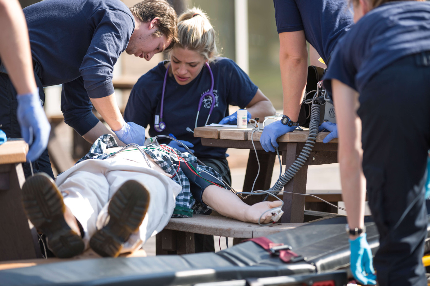 A person is laying on their back while several emergency medical technicians with nitrile gloves, stethescopes and radios are attending to the person. 