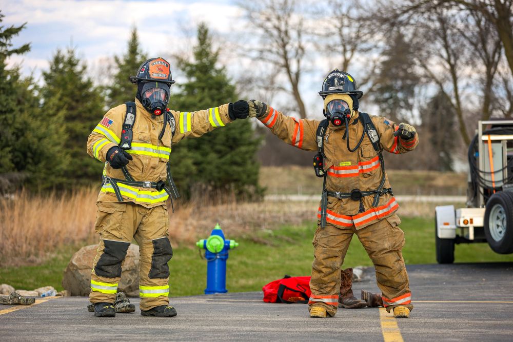 Two fire service students in full fire safety gear bumping fists while standing in an outdoor training area