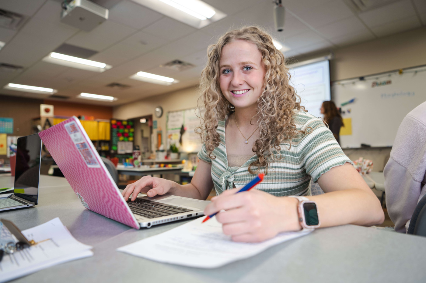 Student in a green striped shirt working on a laptop in a classroom, smiling