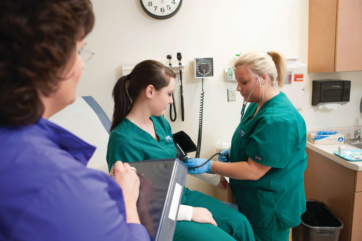 A clinical medical assistant in training takes the blood pressue of a fellow student as an instructor looks on. 