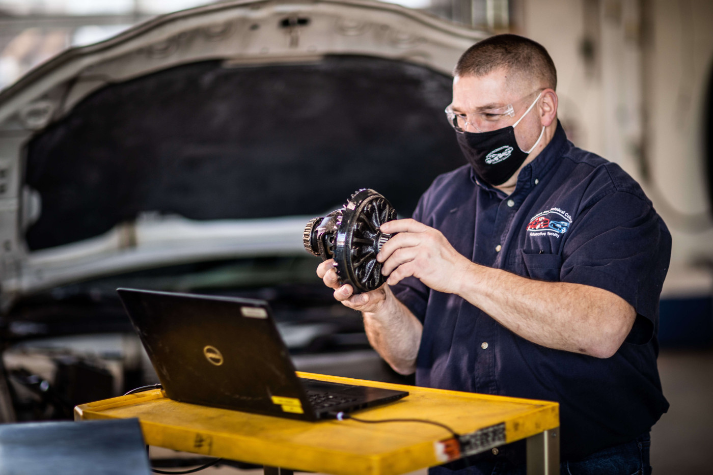 An instructor holds up a part of a motor in an automechanics class. He's removing a part from the hood of a car and demonstrating it.